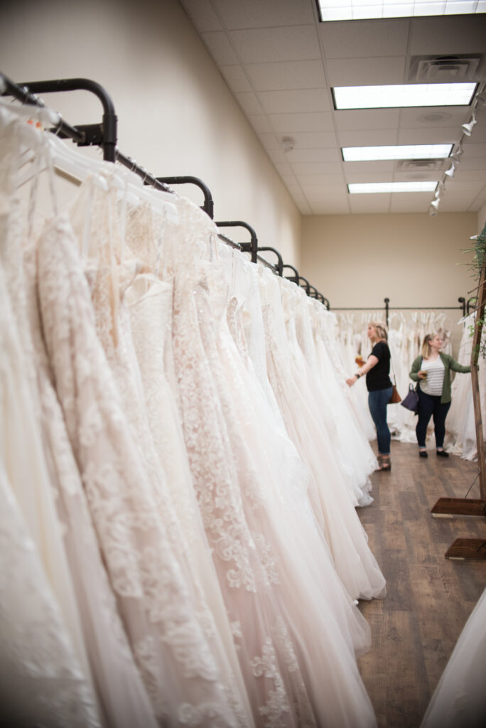 A row of white wedding dresses on racks in a bridal shop, with two women browsing in the background.