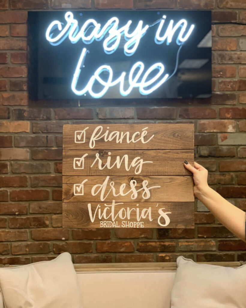 A hand holds a wooden sign with bridal checklist items; a neon "crazy in love" sign is on a brick wall behind.