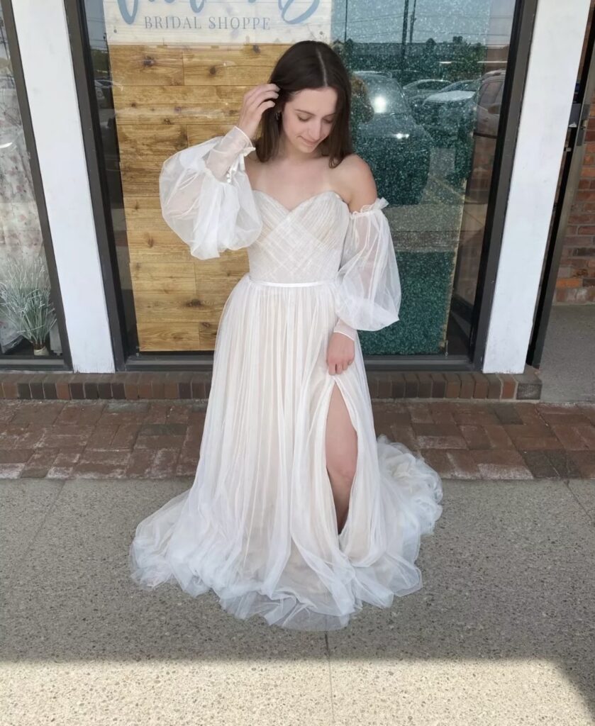 Woman in a white, off-shoulder wedding dress with sheer sleeves and a slit stands outside a bridal shop.