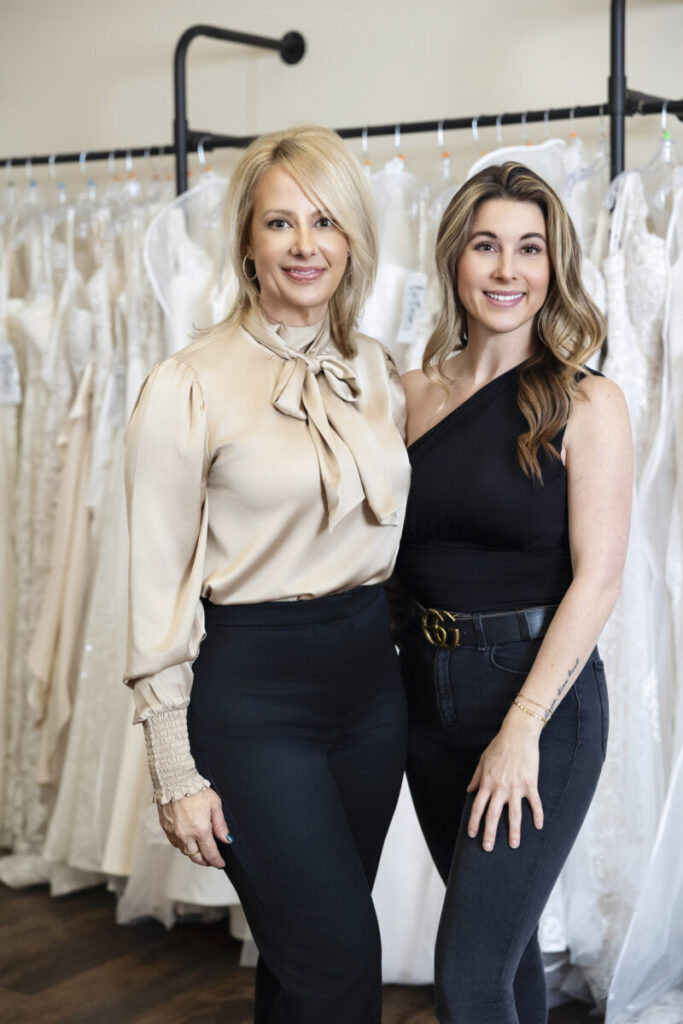 Two women stand side by side in front of a rack of wedding dresses in a bridal shop.