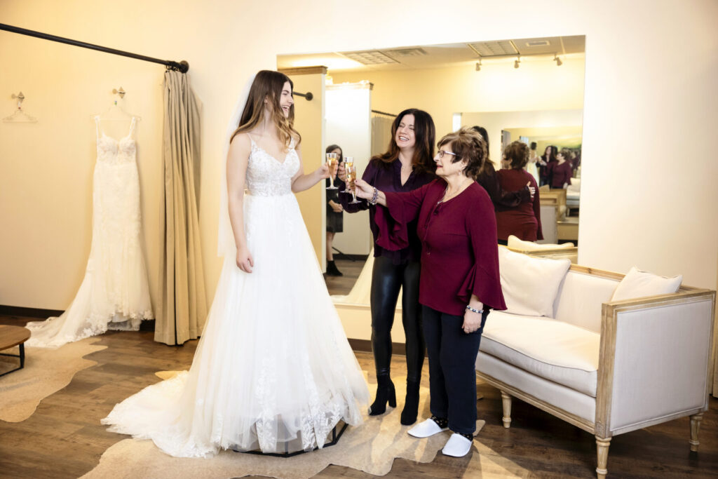 A bride in a wedding dress toasts with two women in a bridal shop fitting room. A dress hangs on the wall nearby.