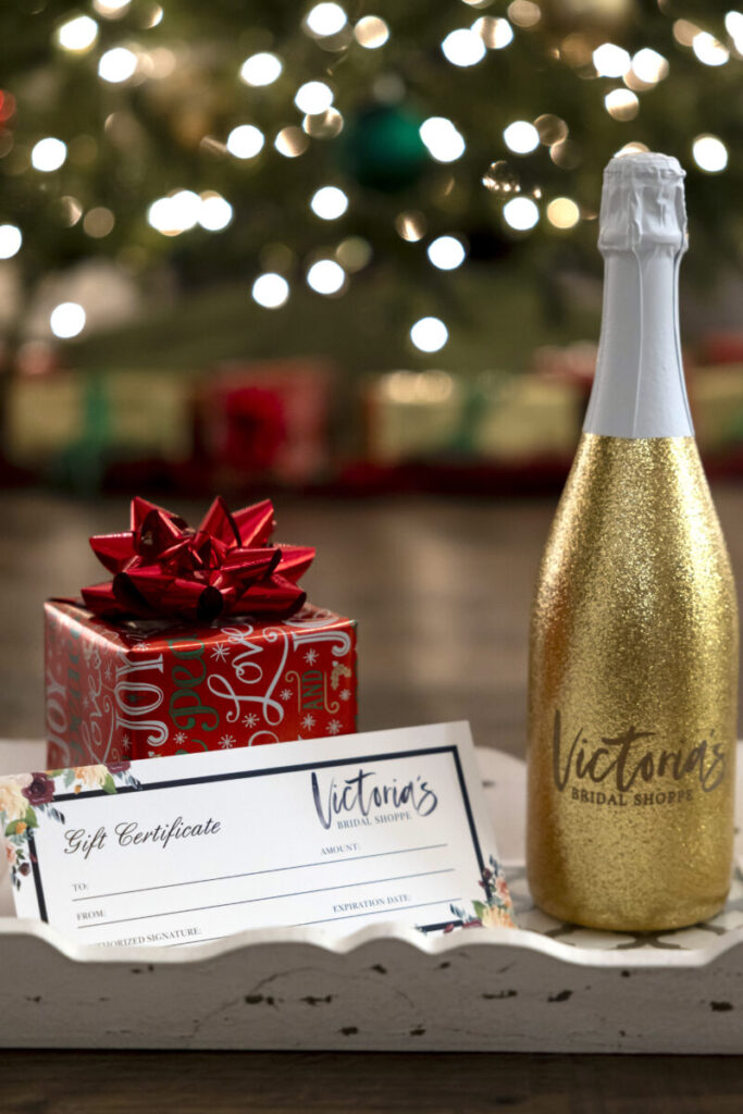 A gold bottle, a red gift box with a bow, and a gift certificate on a tray, with a lit Christmas tree in the background.