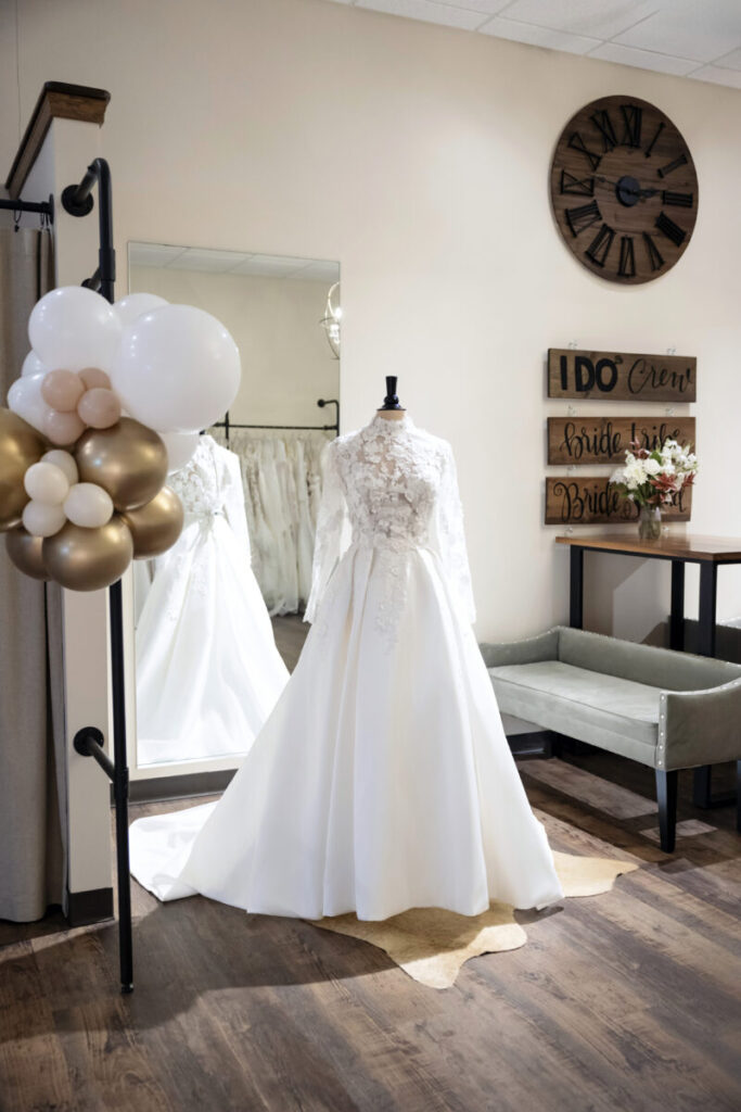 A white lace wedding dress on a mannequin in a bridal shop, with a mirror, bench, and decorative signs in the background.