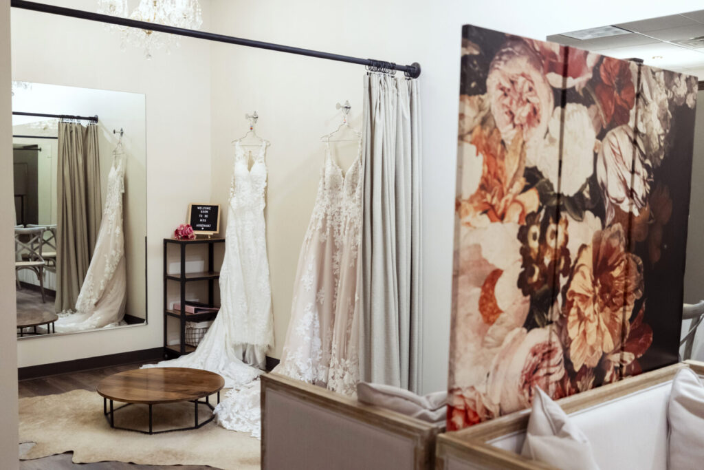 Two wedding dresses on display in a bridal boutique fitting room with a mirror and floral screen.