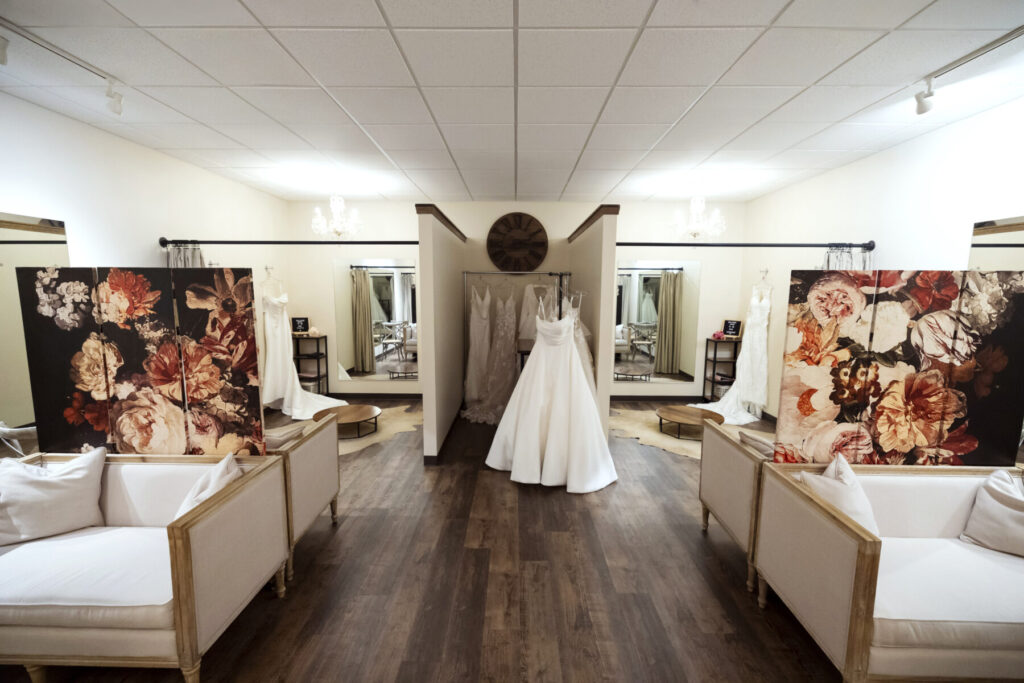 A bridal boutique with wedding dresses on display, floral room dividers, and white seating on a wooden floor.