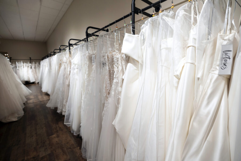A row of white wedding dresses hangs on racks in a bridal shop with a sign marked “Victorian.”.
