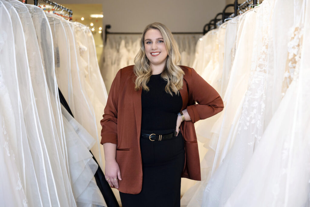 A woman stands smiling among racks of white wedding dresses in a bridal boutique.