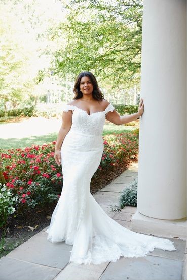 Woman in an off-shoulder white wedding dress stands by a column in a garden with red flowers and greenery.
