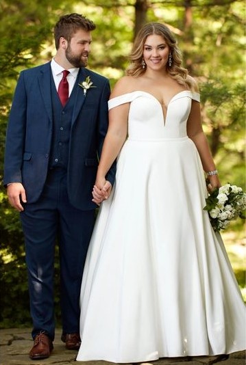 Bride in an off-shoulder white gown holding a bouquet, standing with groom in a blue suit outdoors.
