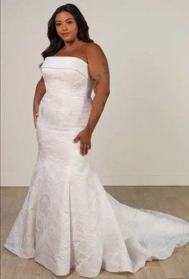 Woman wearing a strapless, fitted white wedding dress with a train, standing on a wooden floor against a plain background.