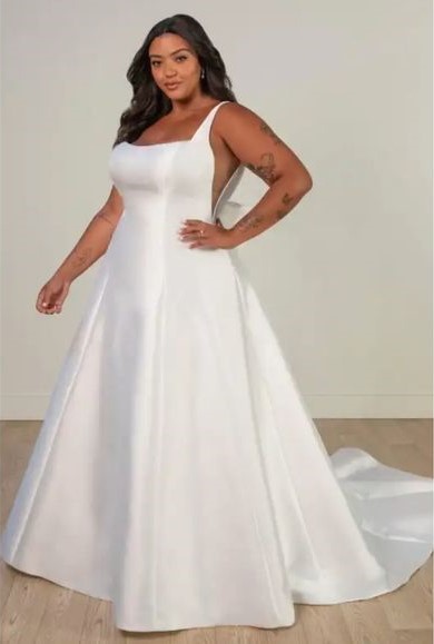 Woman wearing a sleeveless white wedding dress with a fitted bodice and a long, flared train, standing indoors.