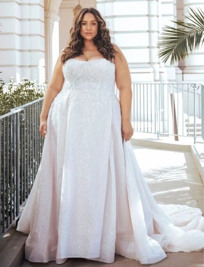 A woman in a white strapless wedding dress stands outdoors near a railing, with sunlight and greenery in the background.