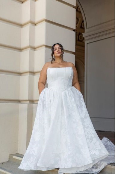 Woman in a strapless white wedding dress stands outside against a cream-colored building with classical architectural details.