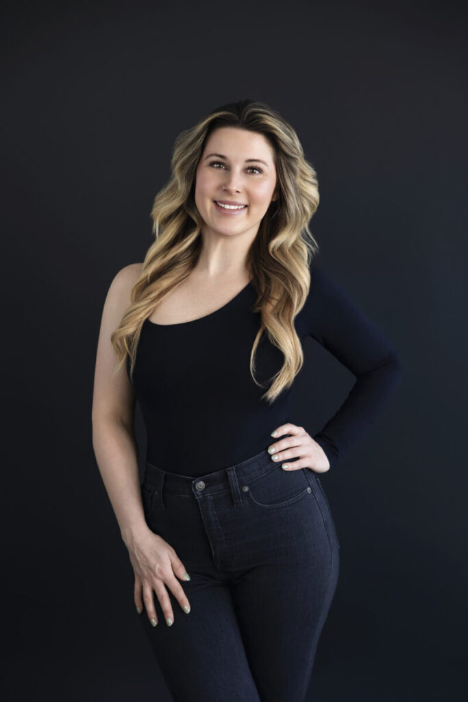 Woman with long blonde hair in a black one-sleeve top and jeans stands against a dark background, smiling at the camera.