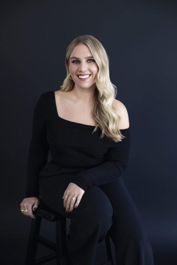 Woman with long blonde hair wearing a black, off-shoulder top, seated on a stool against a dark background.