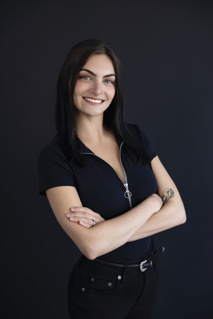 Woman with long dark hair stands against a plain dark background, smiling with arms crossed, wearing a black outfit.
