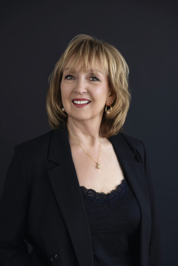 A woman with blonde hair in a black blazer and lace top, smiling against a dark background.