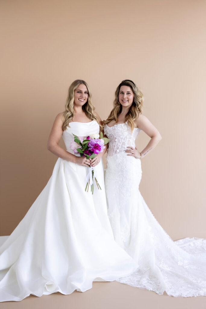 Two women in white wedding dresses stand side by side against a beige background, one holding a bouquet of flowers.
