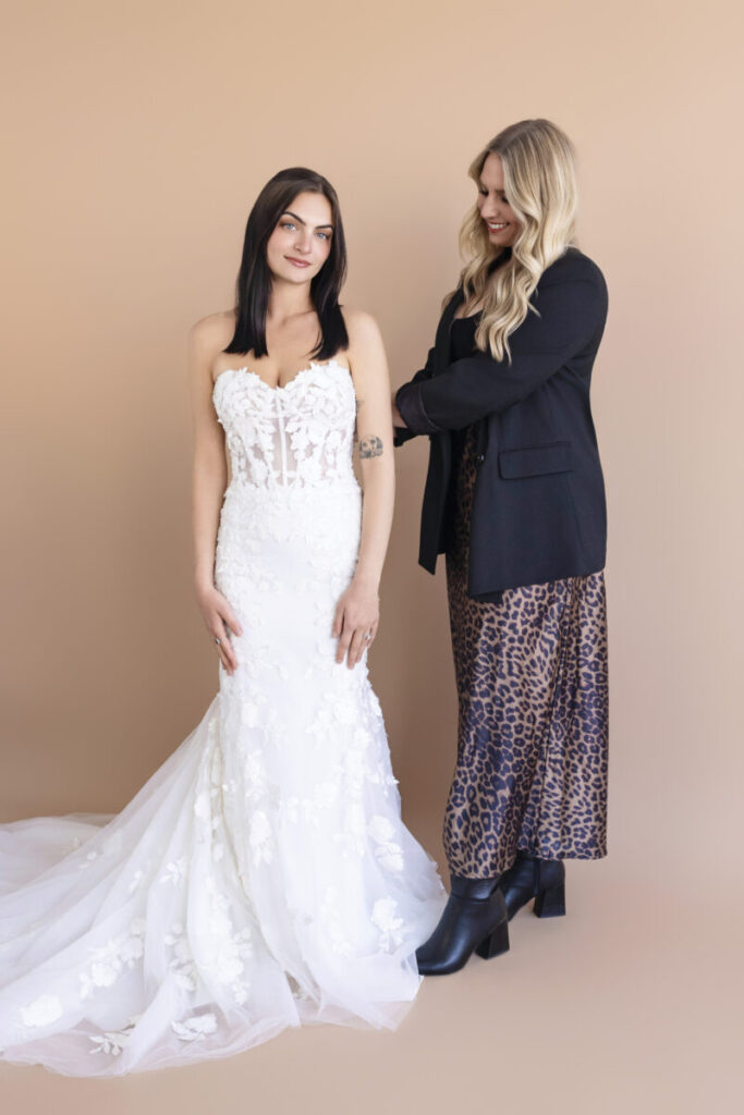 A woman in a white strapless wedding dress stands next to another woman wearing a black blazer and leopard print skirt.