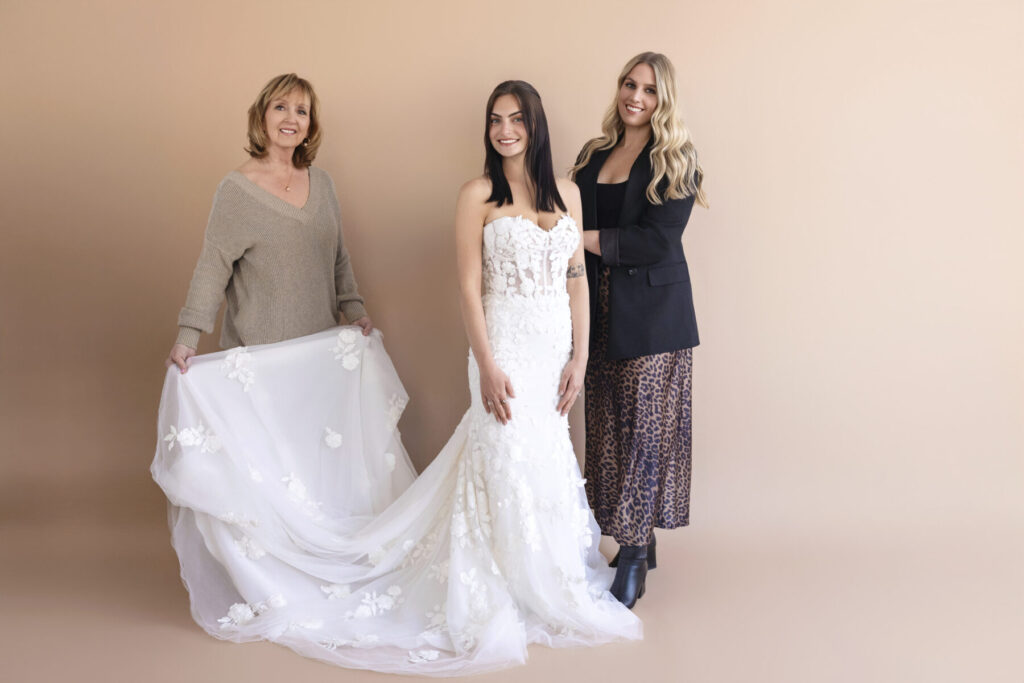 Three women pose together; one wears a wedding dress while the others stand beside and hold the dress’s train.
