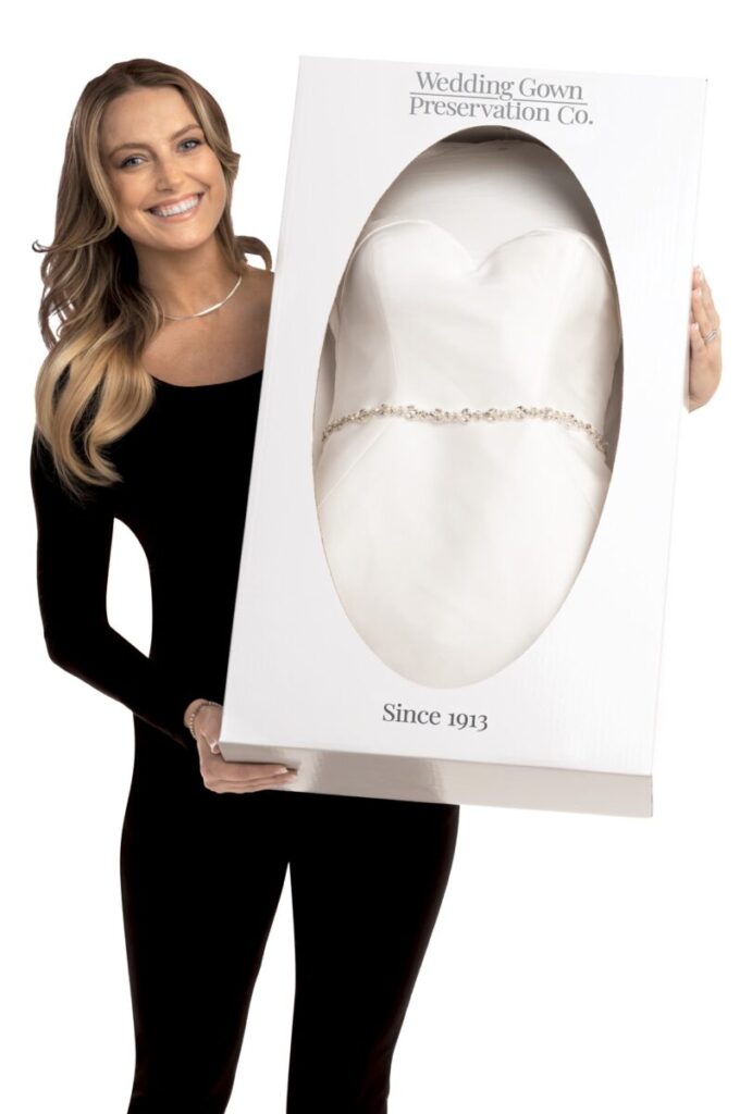 Woman holding a boxed preserved wedding gown from Wedding Gown Preservation Co., visible through a clear window.